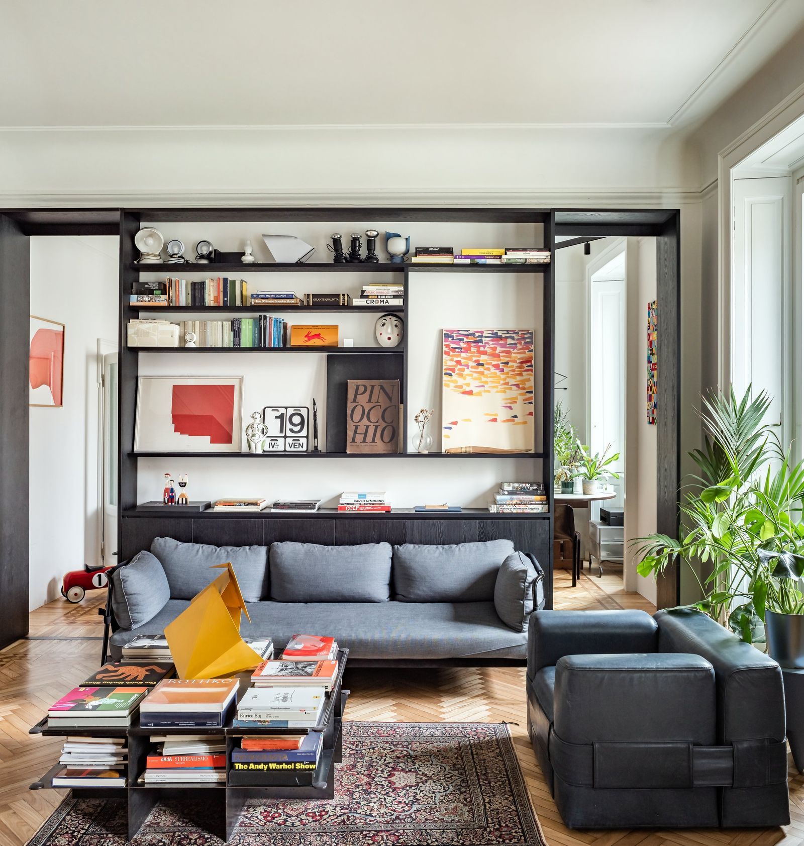 Zona living moderna con libreria a parete, divano Can di Hay, poltrona Cassina 932 di Mario Bellini in pelle blu e tavolino in lamiera d'acciaio NM3.